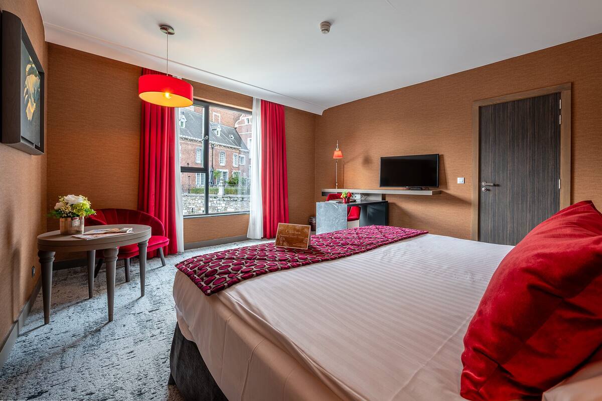 Una habitación de hotel con cama, escritorio, silla, televisor y ventana con cortinas rojas.