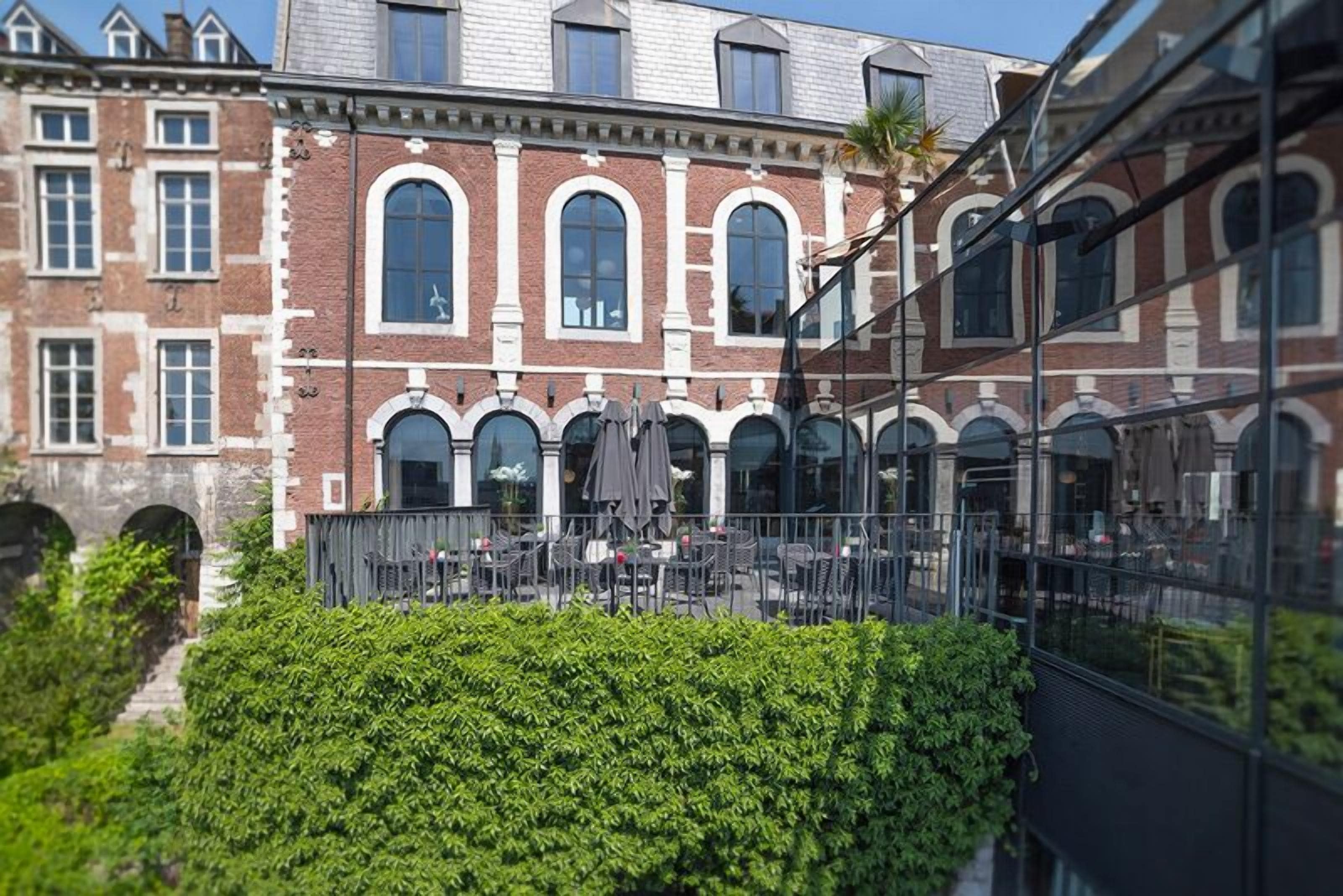 A courtyard with outdoor seating, a hedge, and a building with arched windows and brickwork.