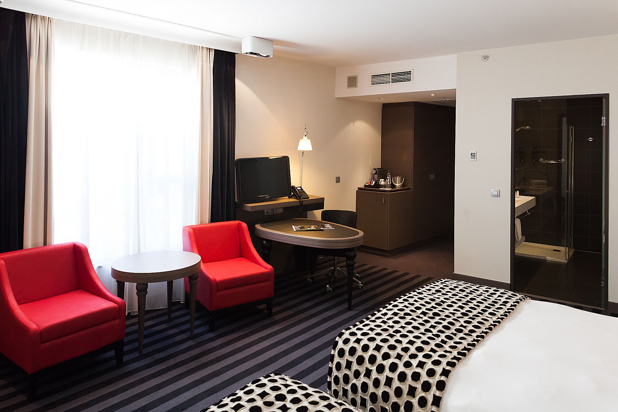 Una habitación de hotel con una cama, un sillón rojo, una mesita, un escritorio con una computadora, un minibar y un baño con puerta de vidrio.