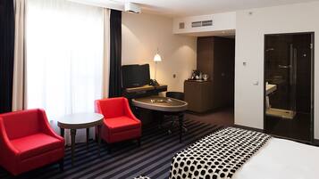Una habitación de hotel con una cama, un sillón rojo, una mesita, un escritorio con una computadora, un minibar y un baño con puerta de vidrio.