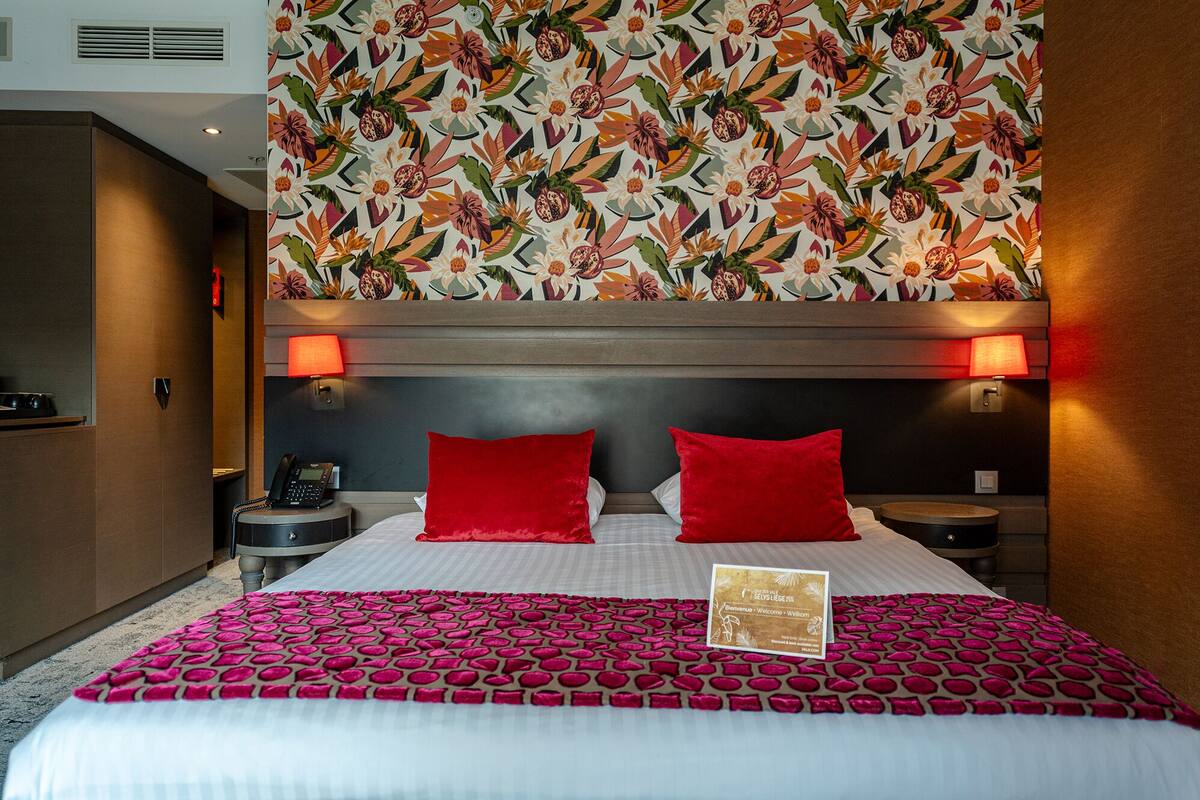 Habitación de hotel con una cama grande, dos almohadas rojas, una mesita de noche con teléfono y una pared con estampado floral.