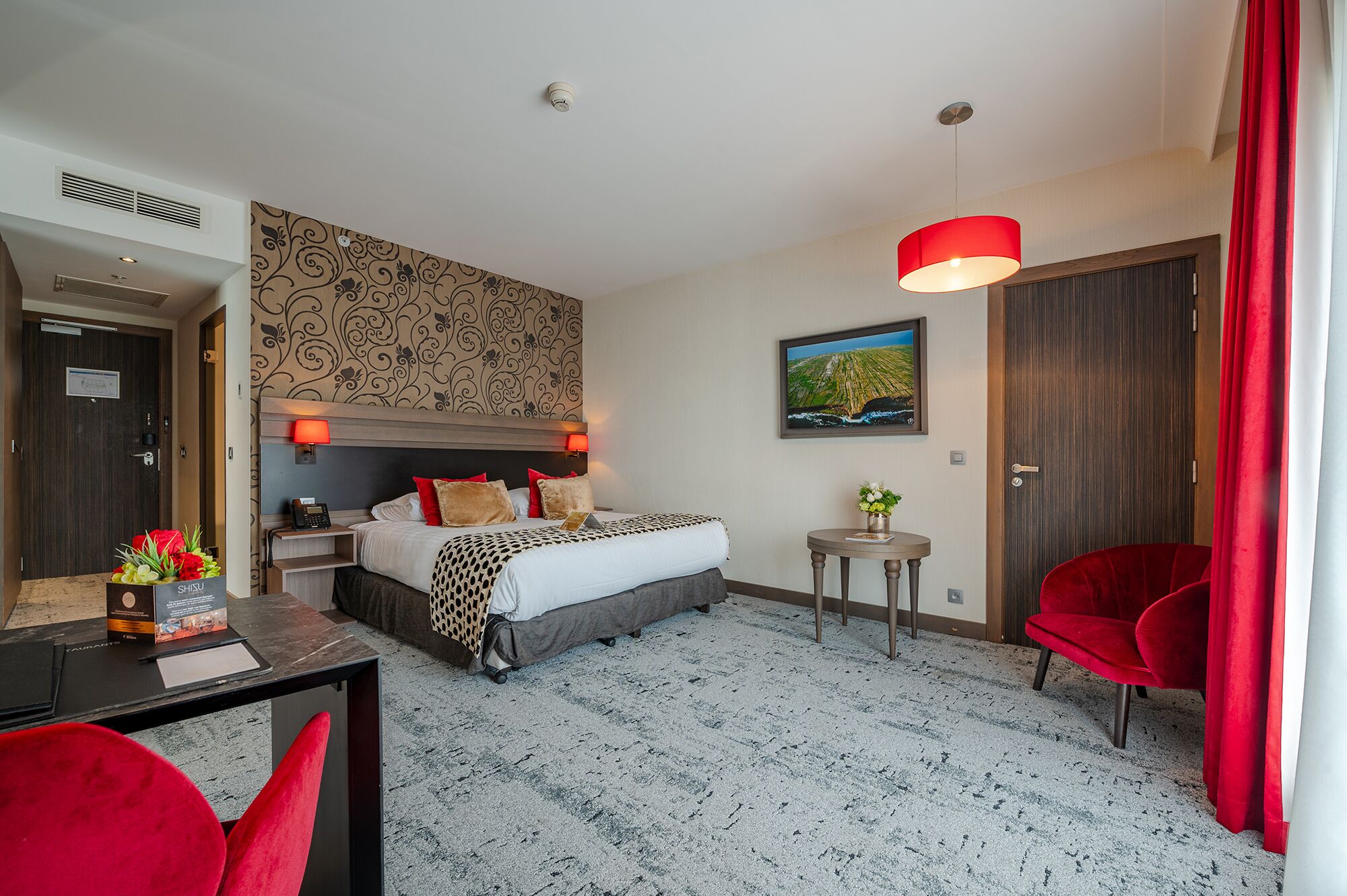 A hotel room with a bed, a desk with a chair, a small table with a vase, and a red chair.