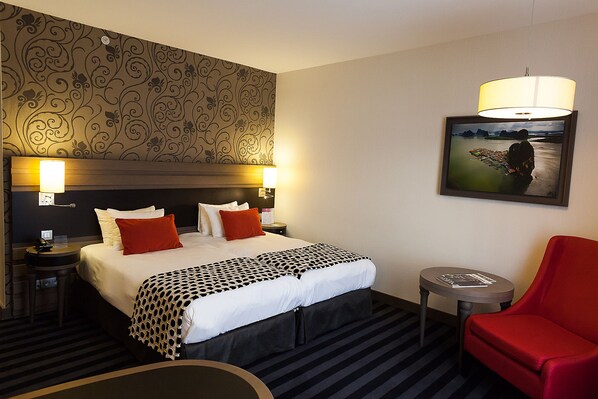 Une chambre d’hôtel avec un grand lit, deux lampes de chevet, un fauteuil rouge, une petite table ronde et un tableau encadré au mur.