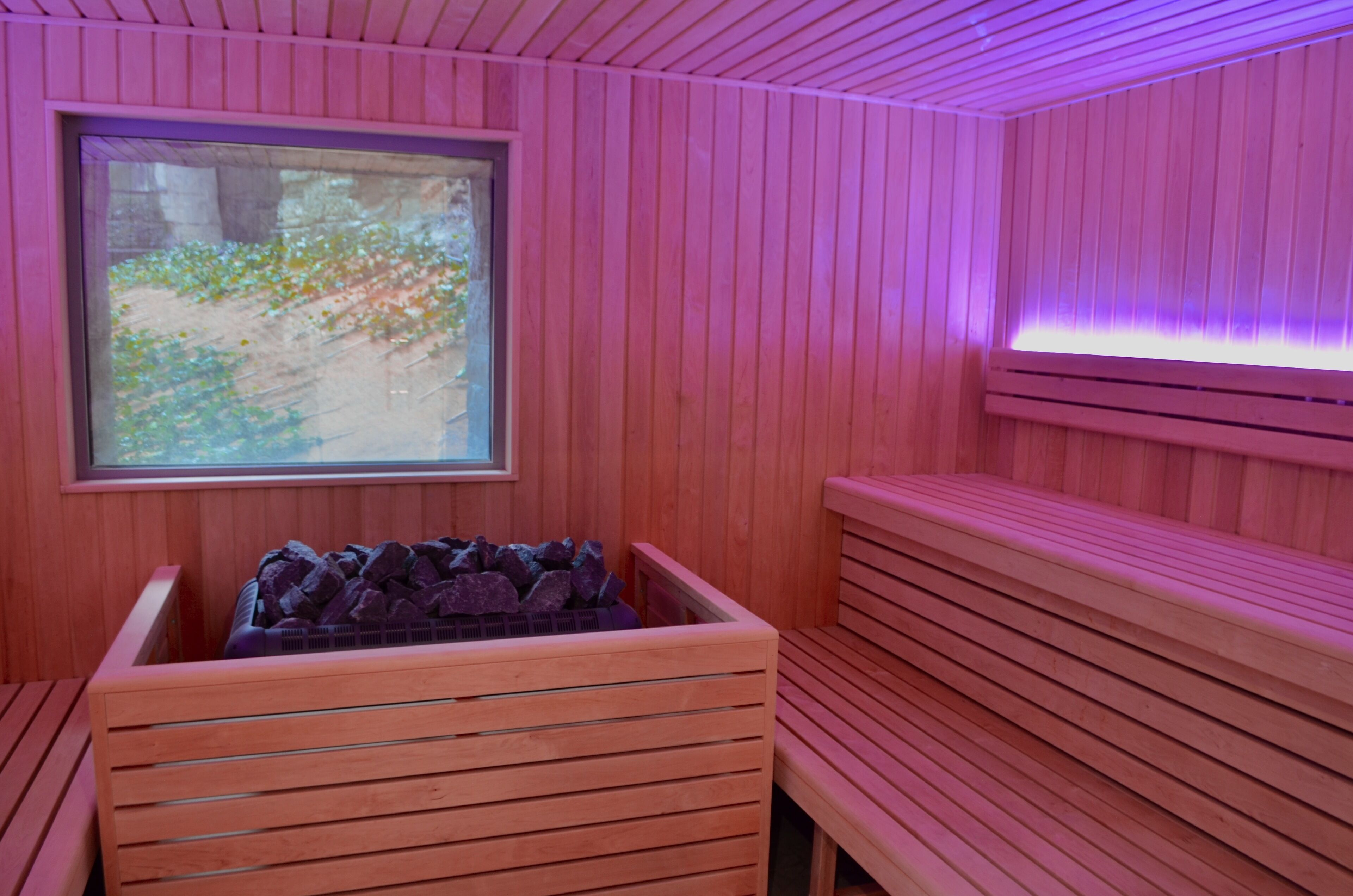 Un sauna con bancos de madera, una ventana con vista a la vegetación y un banco de piedra con rocas.