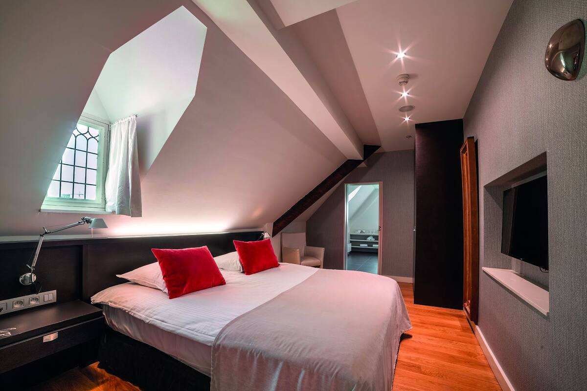 Un dormitorio moderno con tragaluz, una cama con almohadas rojas, una mesita de noche y un televisor montado en la pared.
