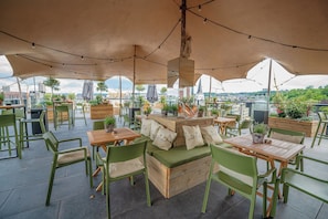 An outdoor seating area with tables and chairs, a central wooden bench, and potted plants. - Van der Valk Sélys Liège Hotel & Spa (Liege)