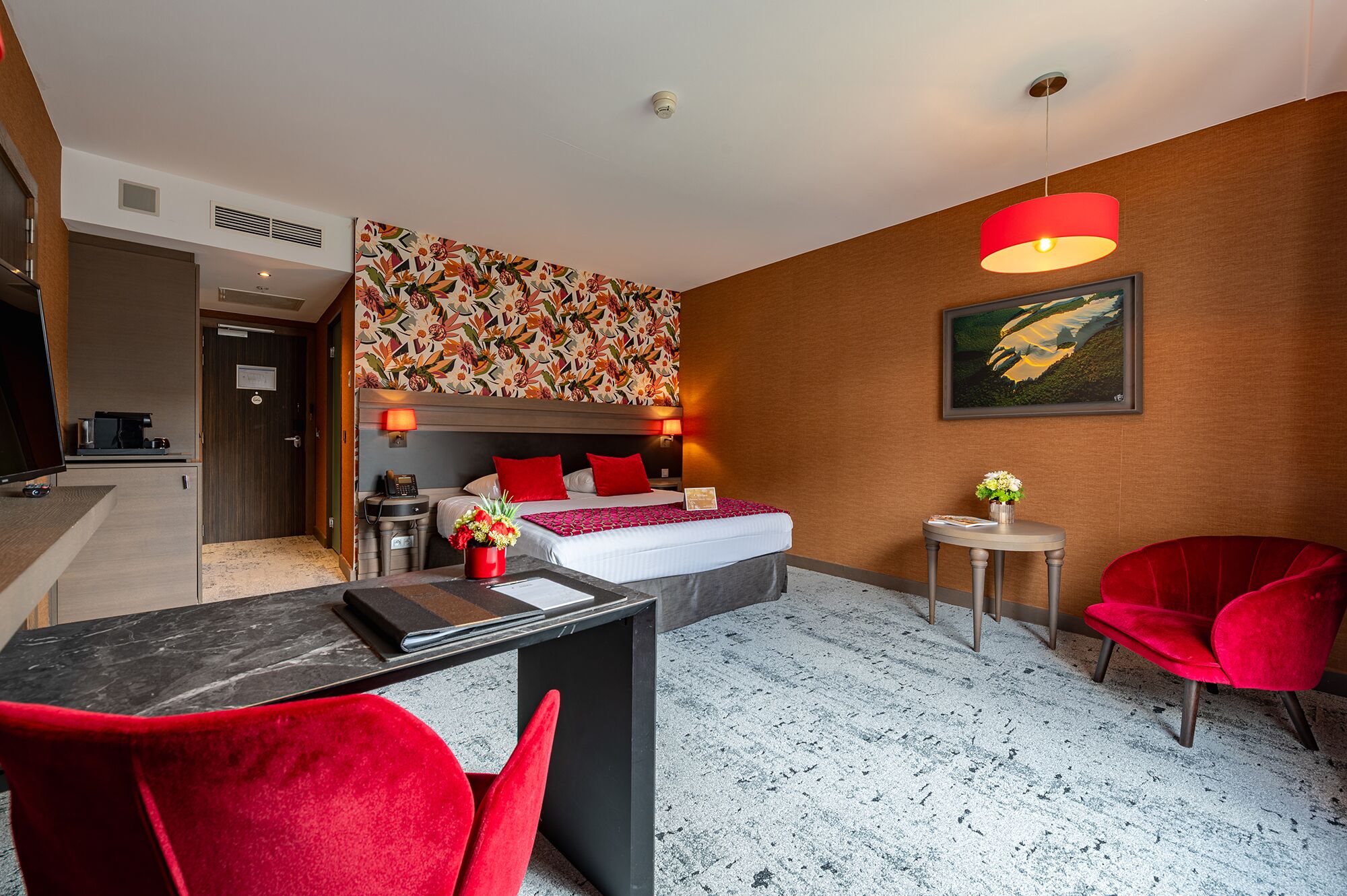 A hotel room with a bed, bedside lamps, a desk with a chair, a small table with a vase, and a red chair.