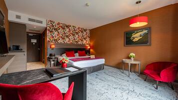 Habitación de hotel con cama, lámparas de noche, un escritorio con silla, una mesita con un florero y una silla roja.