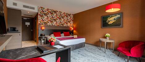 A hotel room with a bed, bedside lamps, a desk with a chair, a small table with a vase, and a red chair.