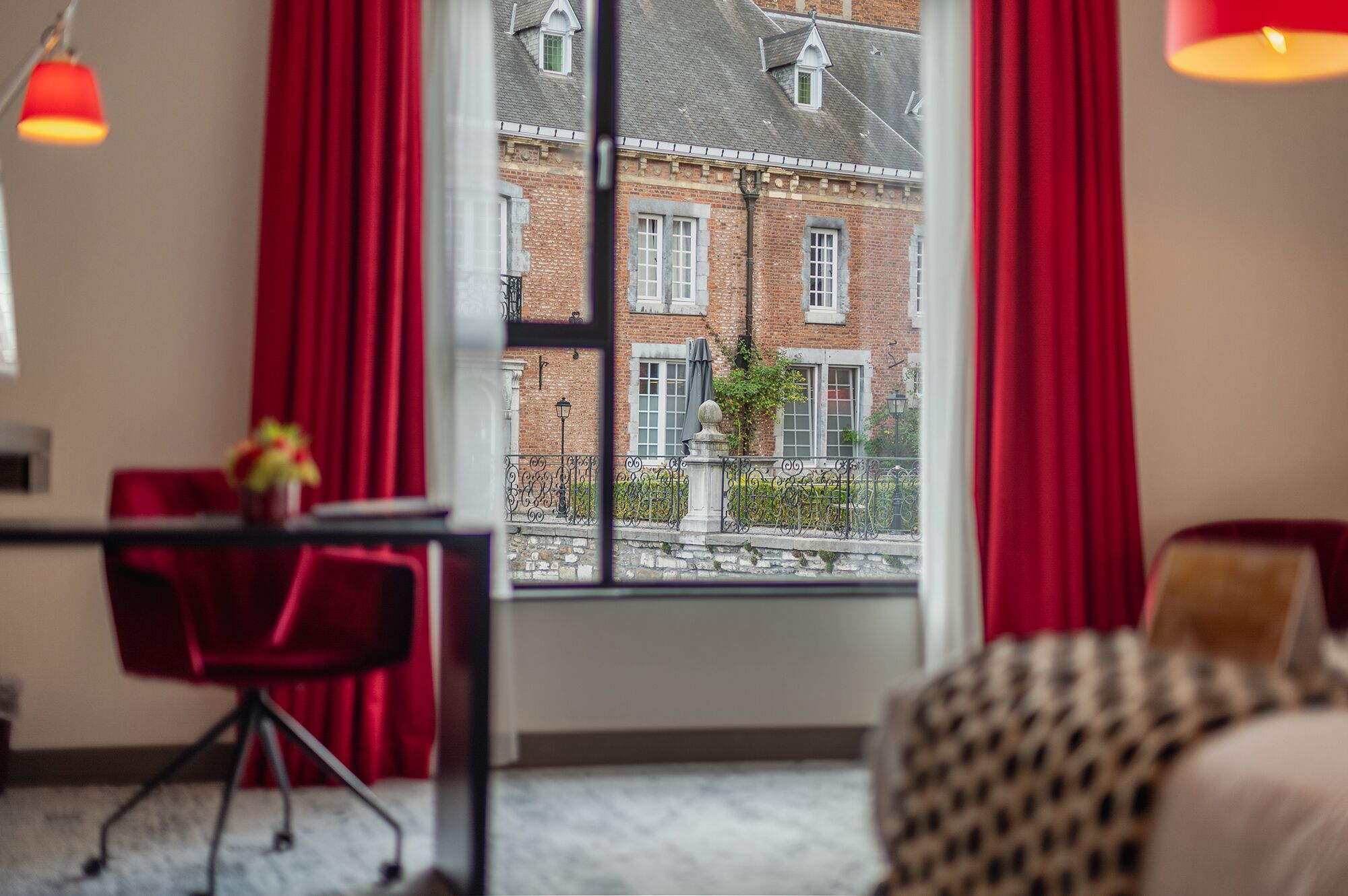 Een kamer met een groot raam, rode gordijnen, een eettafel met stoelen en uitzicht op een bakstenen gebouw.