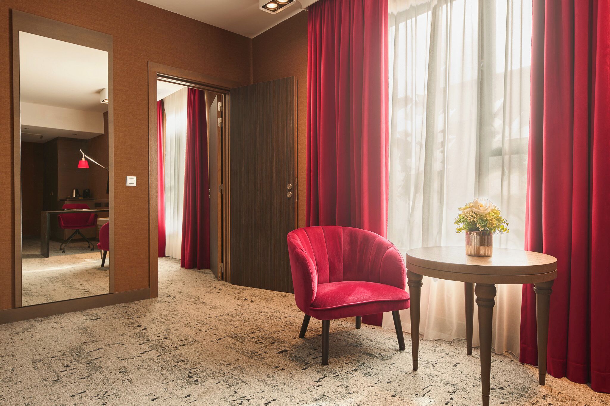 Ein Hotelzimmer mit einem roten Sessel, einem runden Tisch mit Blumenvase und einem großen Spiegel, der einen Schreibtisch und Stuhl reflektiert.