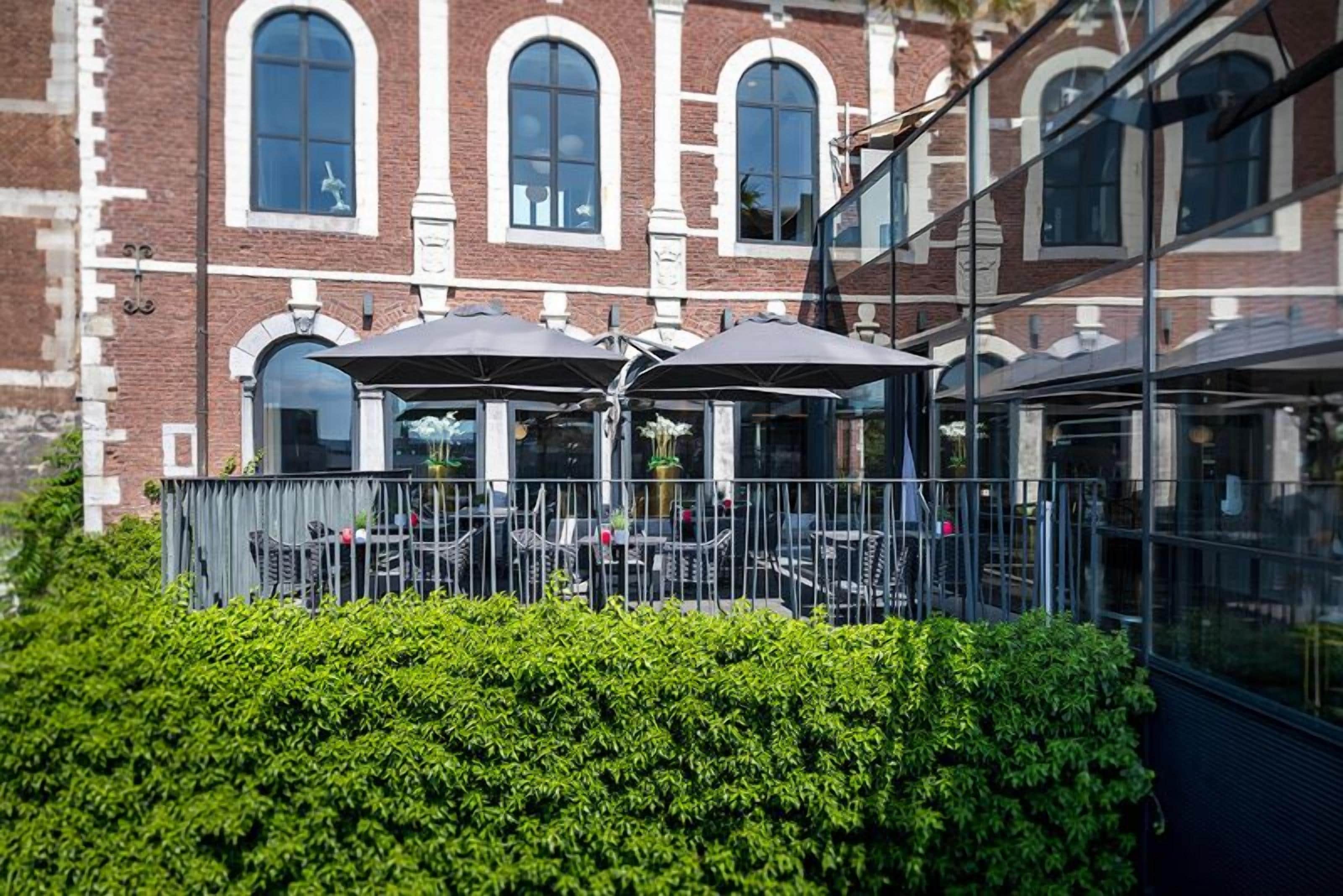 Un patio con mesas al aire libre, un edificio de ladrillo con ventanas de arco y un cerramiento de vidrio.