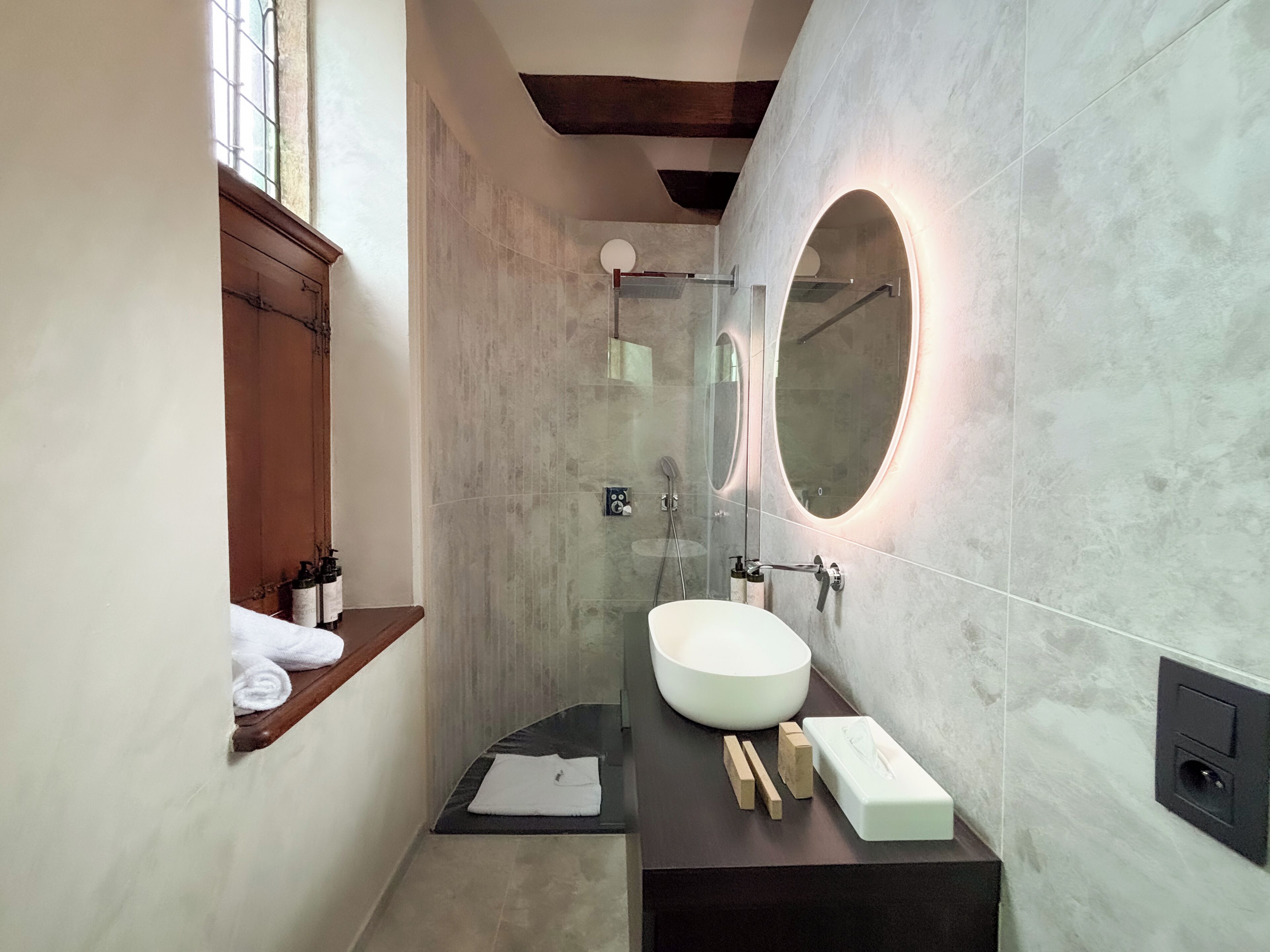 A modern bathroom with a large round mirror, a white sink, and a wooden shelf with a towel.