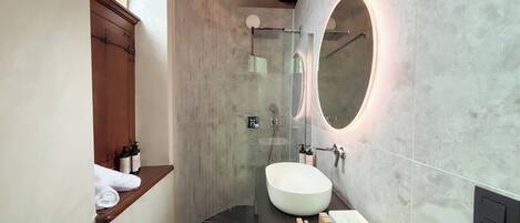 A modern bathroom with a large round mirror, a white sink, and a wooden shelf with a towel.