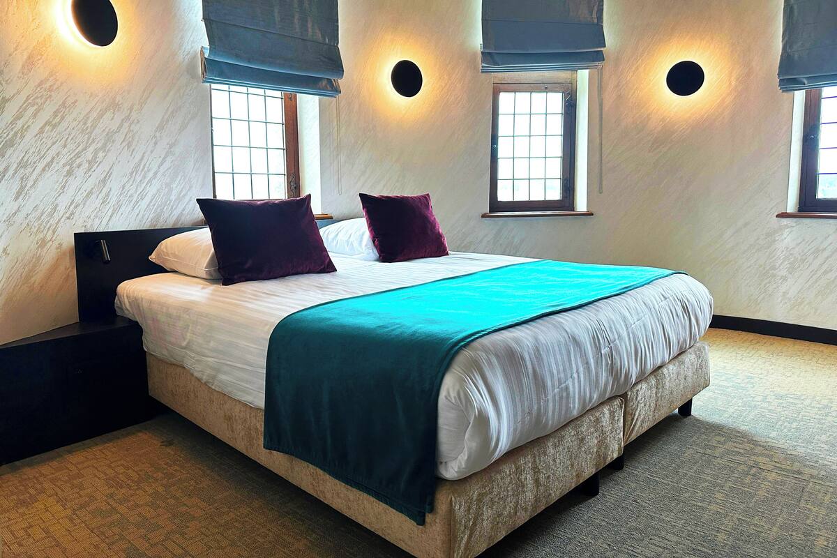 Un dormitorio con una cama grande, dos mesitas de noche, una ventana con persianas y un aplique de pared.