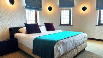 A bedroom with a large bed, two nightstands, a window with blinds, and a wall-mounted light fixture.