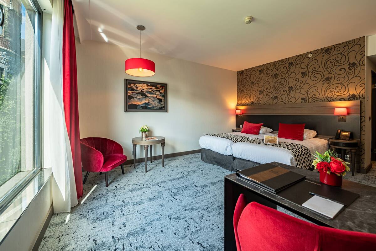 Habitación de hotel con cama, escritorio, silla, una mesita y ventana con cortinas.