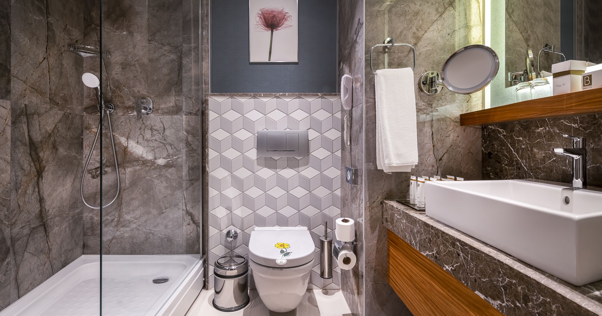 A modern bathroom with a glass shower enclosure, a toilet with a flower decal, a toilet paper holder, and a sink with a mirror above it.