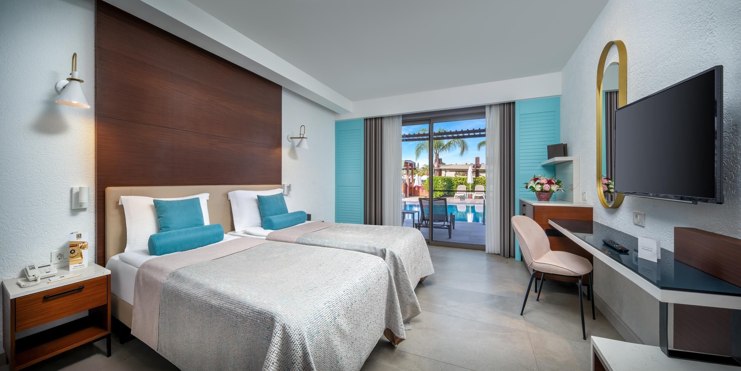 A hotel room with two beds, a desk, a TV, and a view of a pool and palm trees.