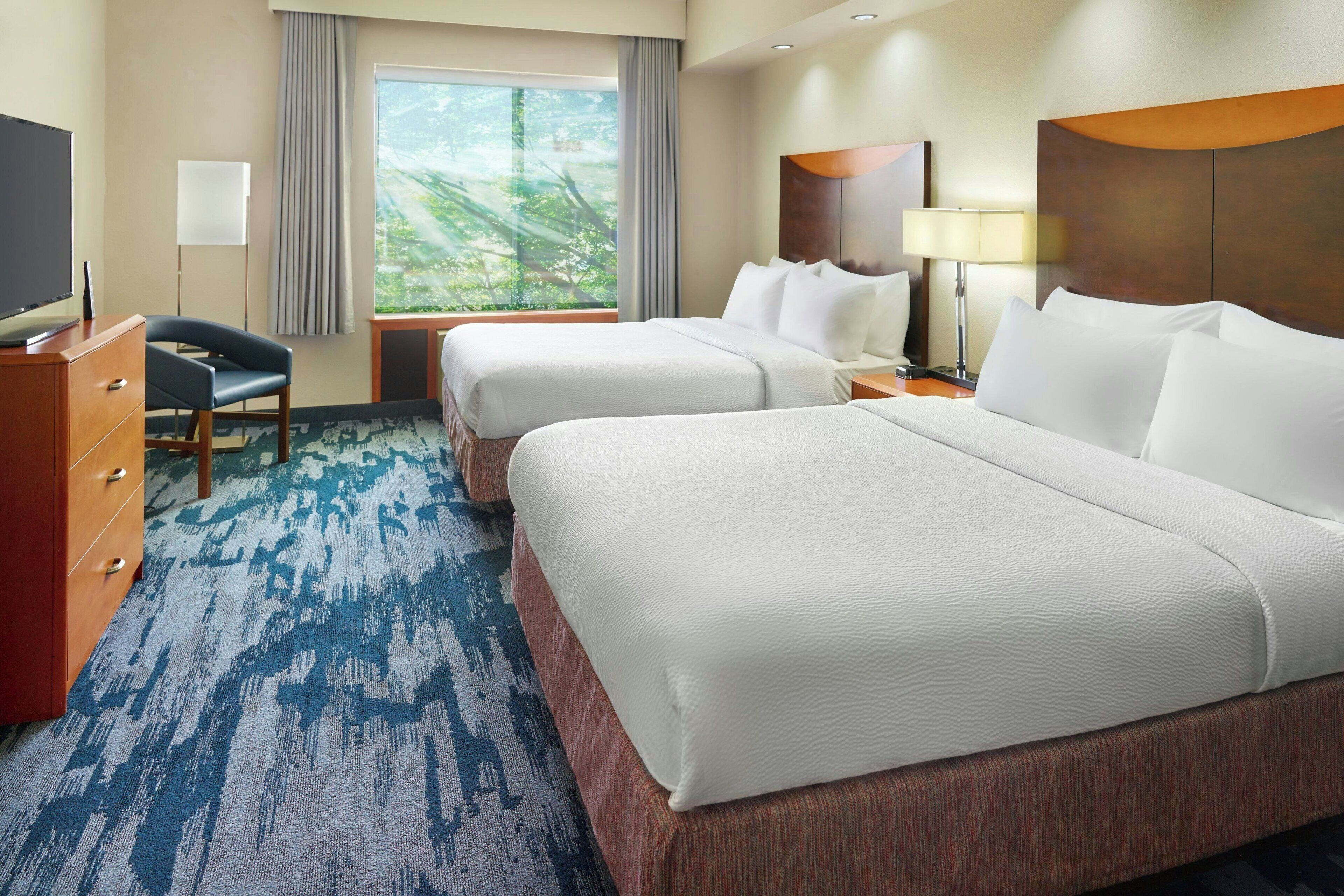 A hotel room with two beds, a TV, a desk, and a large window with a view of greenery.