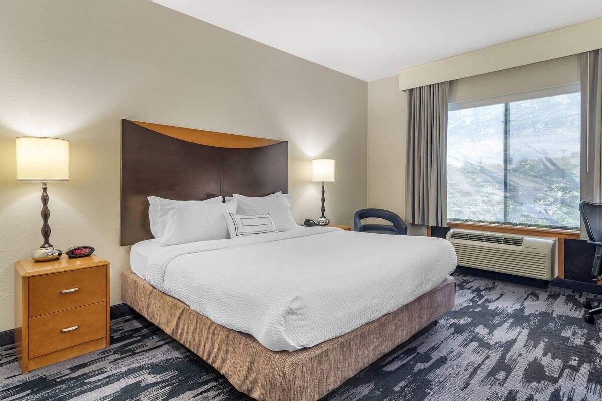 A hotel room with a large bed, two bedside lamps, a desk with a chair, and a window with a view of trees.