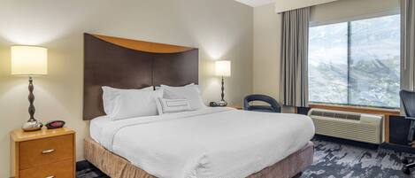 A hotel room with a large bed, two bedside lamps, a desk with a chair, and a window with a view of trees.