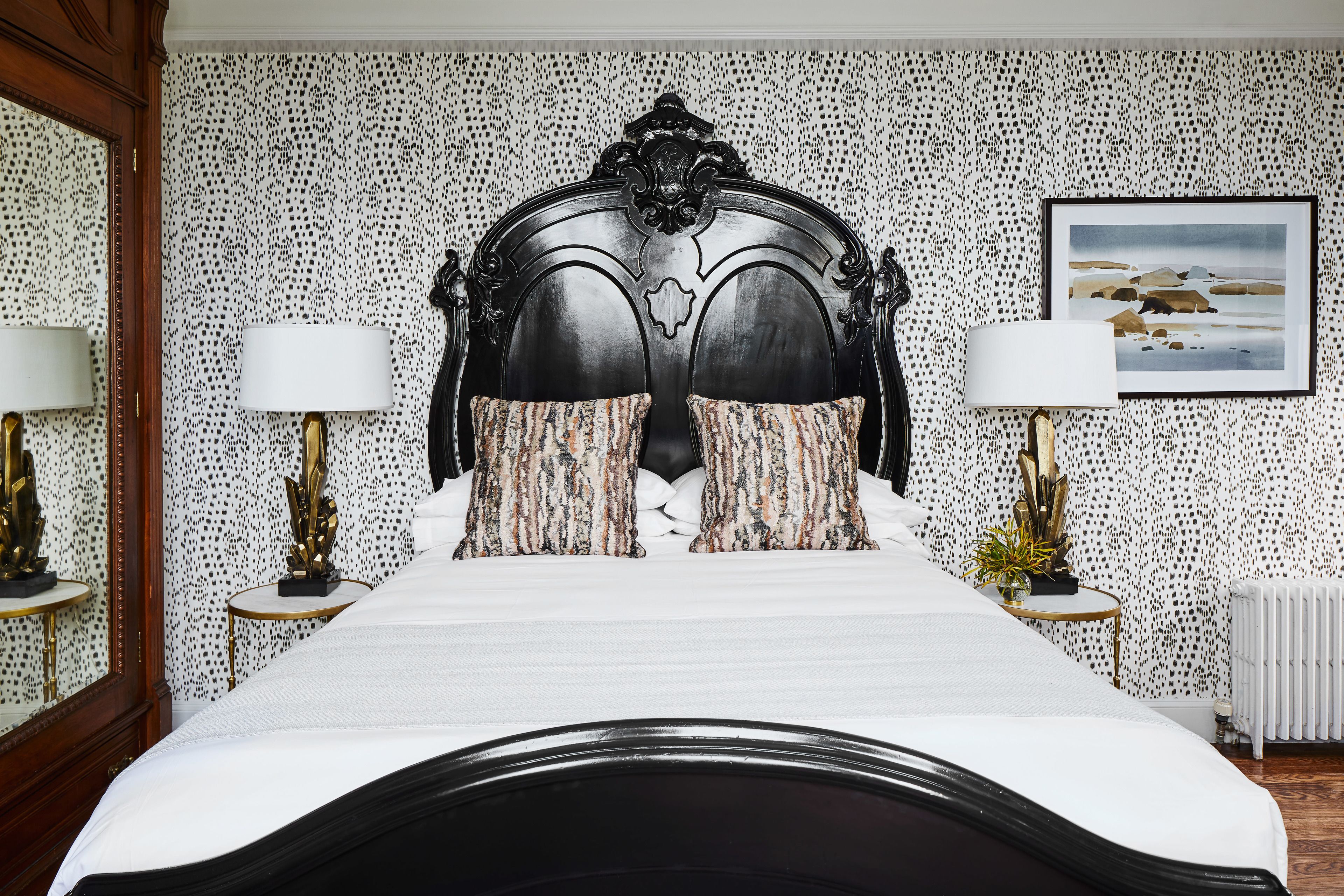 A bed with a dark headboard, two bedside lamps, and patterned pillows. A framed picture is on the wall above the bed.