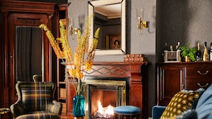 Una sala de estar acogedora con chimenea, un sillón escocés, un reposapiés azul y un jarrón con flores secas.