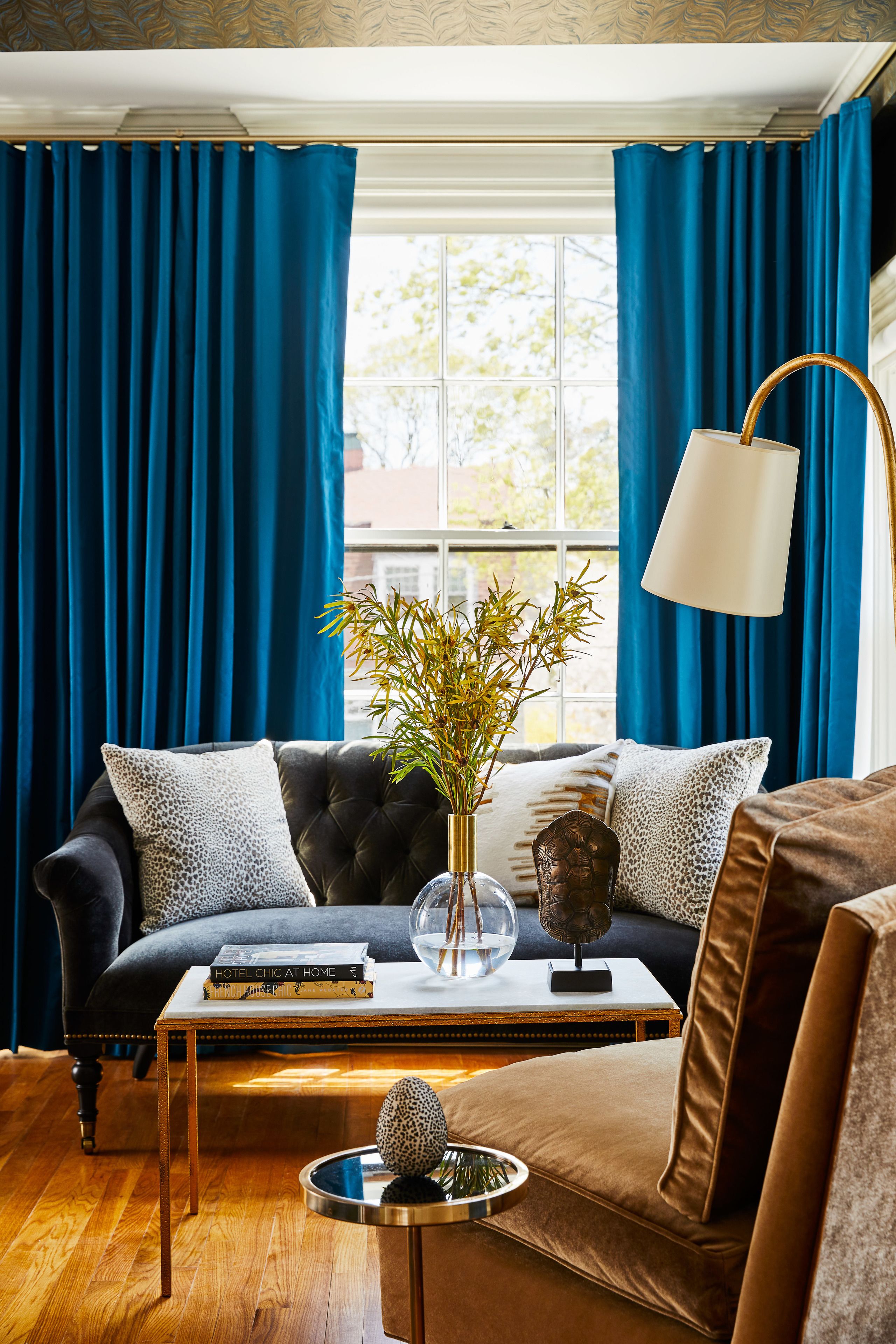 A living room with a dark blue sofa, a white coffee table, and a lamp. There is a window with blue curtains and a view of trees outside.