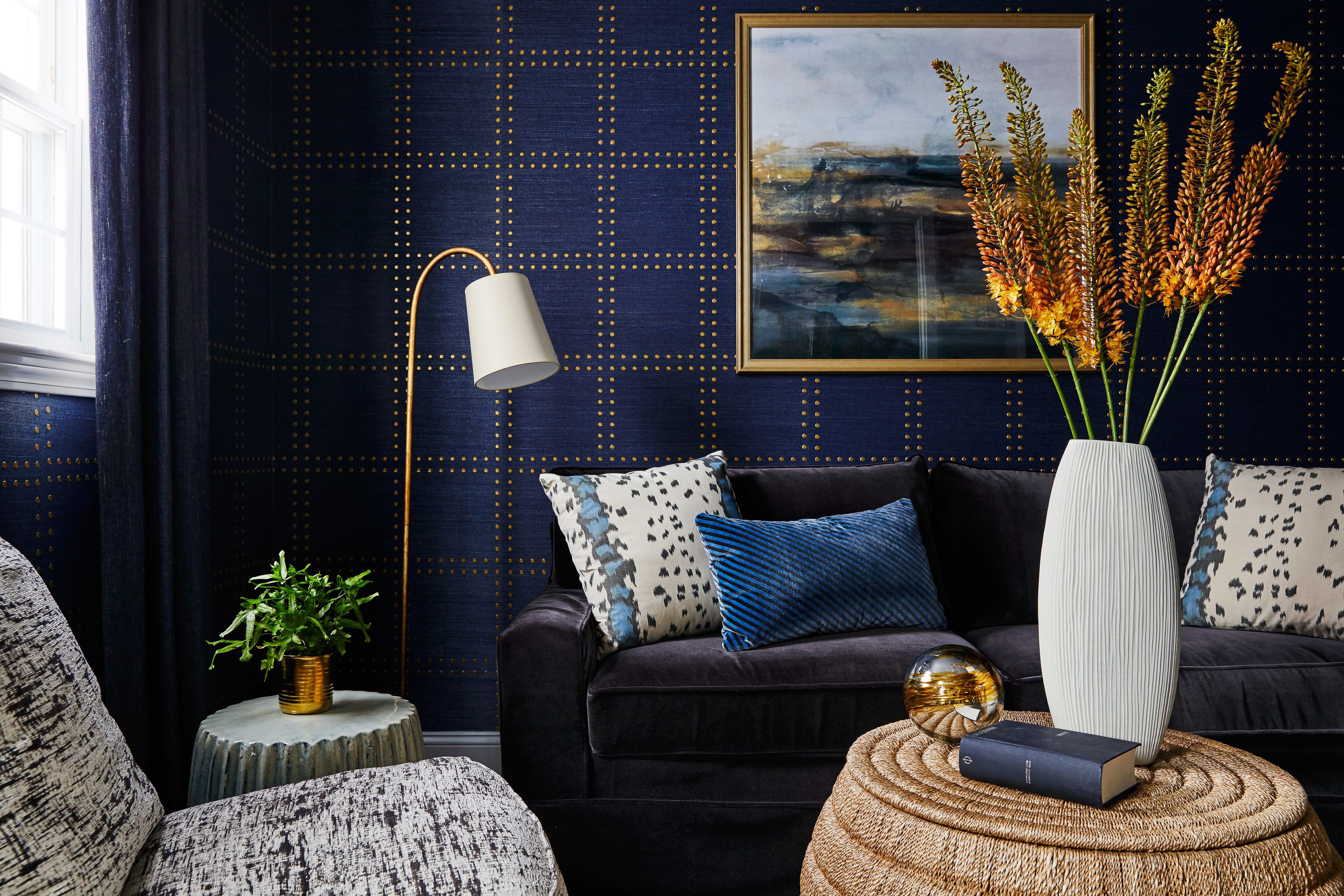 A living room with a dark blue patterned wall, a black sofa with patterned cushions, a round wooden side table, a white vase with plants, a floor lamp, and a framed artwork.