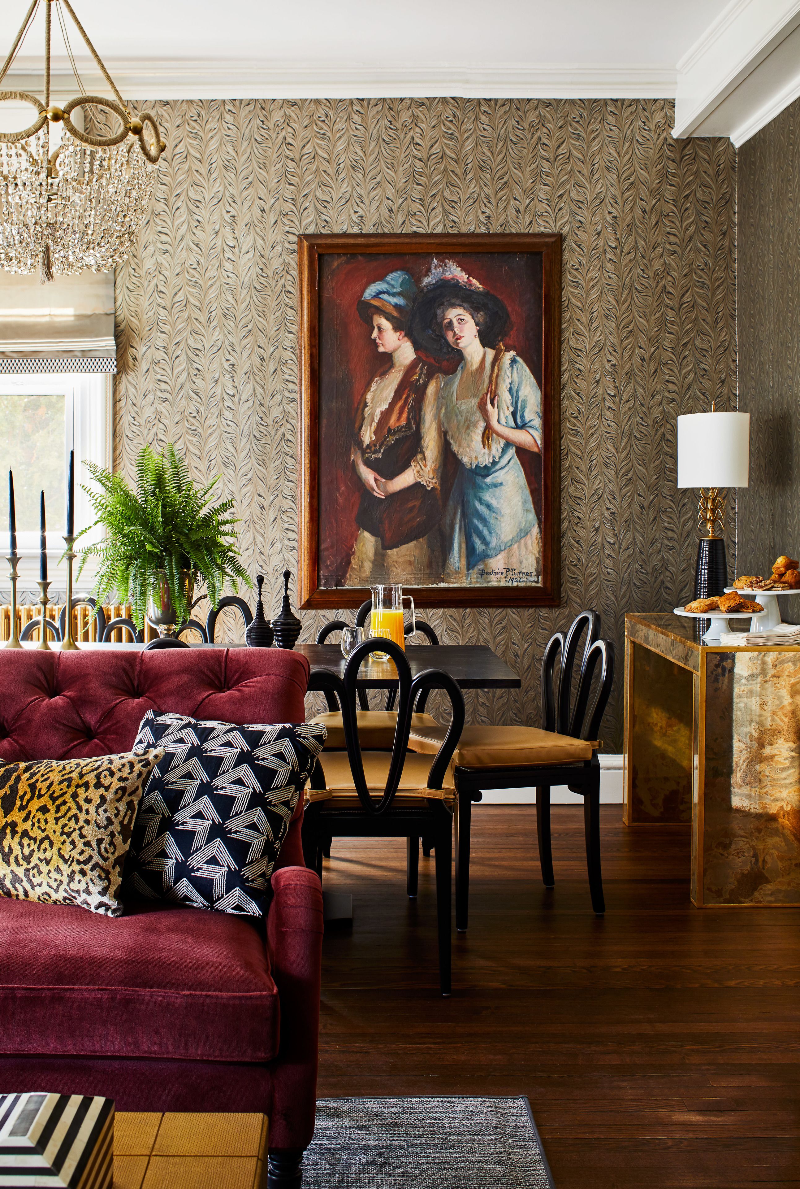 A dining area with a red sofa, patterned wallpaper, a chandelier, and a painting on the wall.