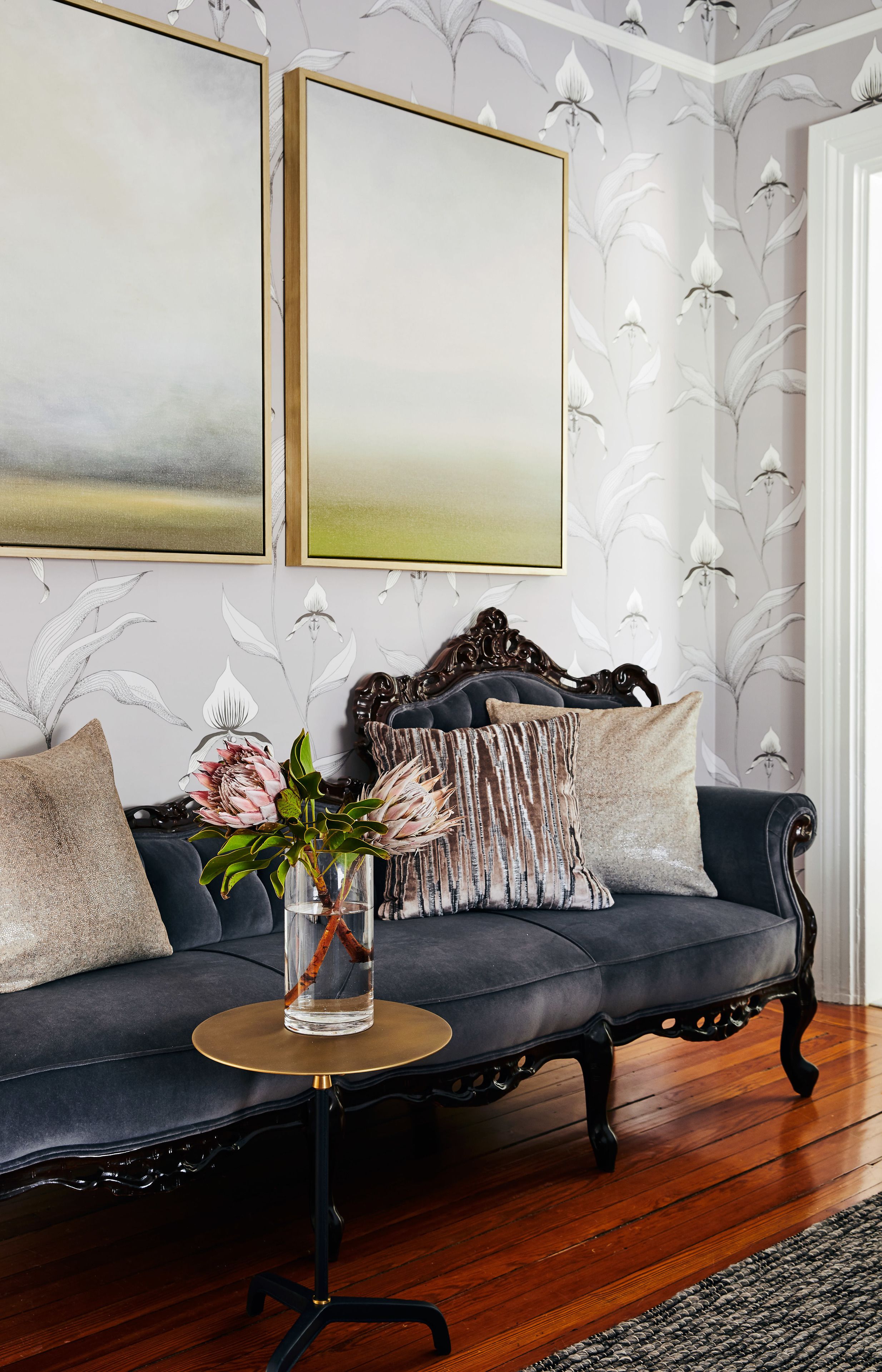 Ein dunkelblaues Sofa mit dekorativer Kopfteil, ein runder Tisch mit Blumenvase und floraler Wandtapete.