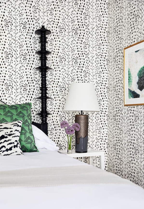 A bedroom with a patterned wall, a bed with patterned pillows, a bedside table with a lamp, and a framed picture on the wall.