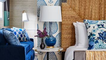 A room with a blue sofa, a bed with a woven headboard, a bedside table with a lamp, and patterned wallpaper.