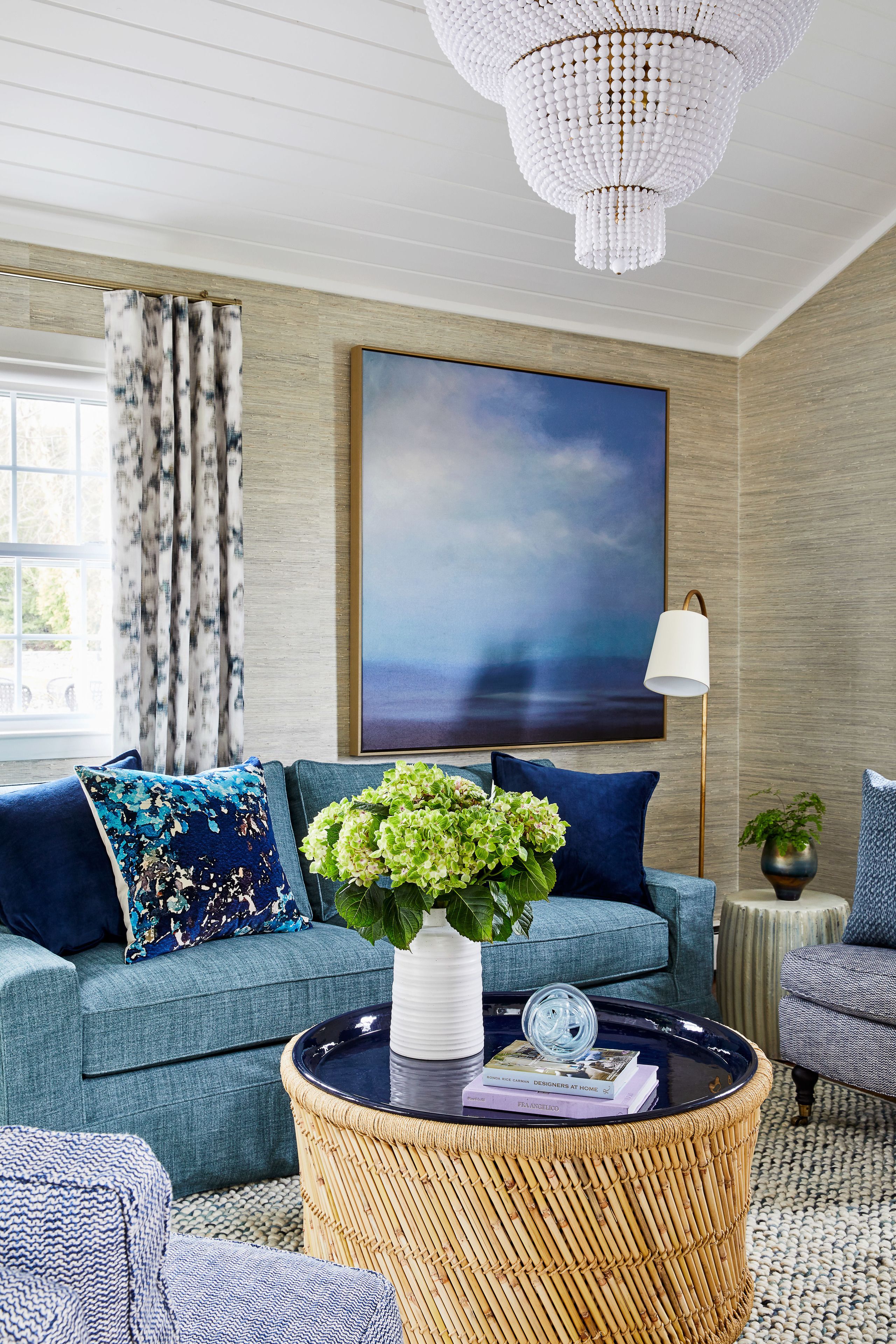 A modern living room with a blue sofa, a round wicker coffee table, and a large painting on the wall.