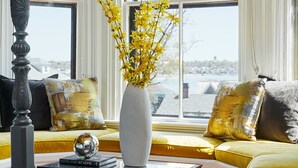 Una habitación con un ventanal grande, un sofá con cojines amarillos, un jarrón blanco con flores amarillas y una mesa de centro de madera.