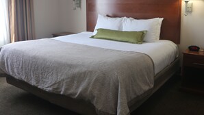 A neatly made bed with a patterned bedspread, a green pillow, and white pillows. There is a wooden headboard, a nightstand with a lamp, and a digital clock displaying the time.