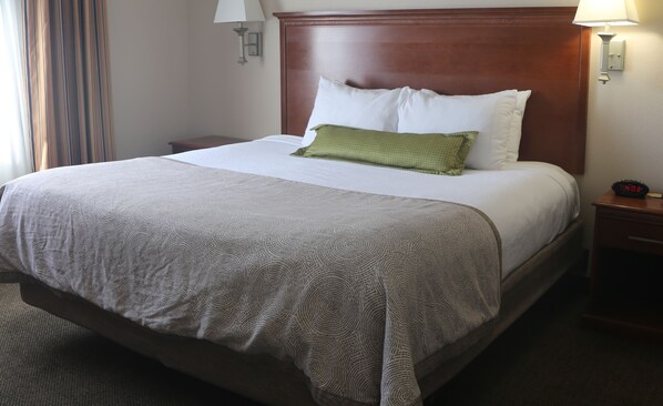 A neatly made bed with a patterned bedspread, a green pillow, and white pillows. There is a wooden headboard, a nightstand with a lamp, and a digital clock displaying the time.