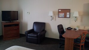 A hotel room with a flat-screen TV, a leather chair, a wooden table with two chairs, and a bulletin board with papers and a map.