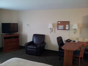 A hotel room with a flat-screen TV, a leather chair, a wooden table with two chairs, and a bulletin board with papers and a map.