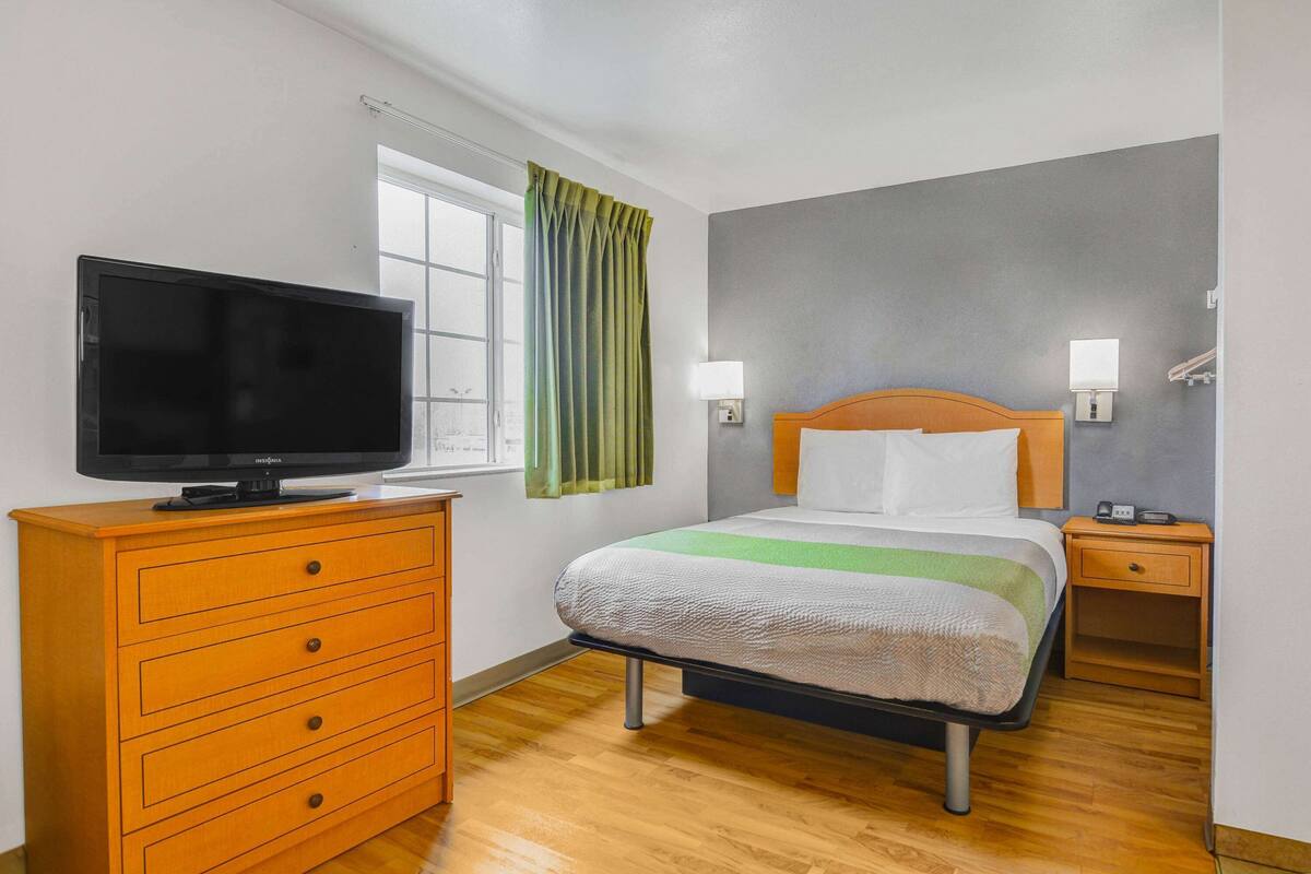 Quarto de hotel com cama, televisão num penteado, janela com cortinas e mesa de cabeceira com abajur.