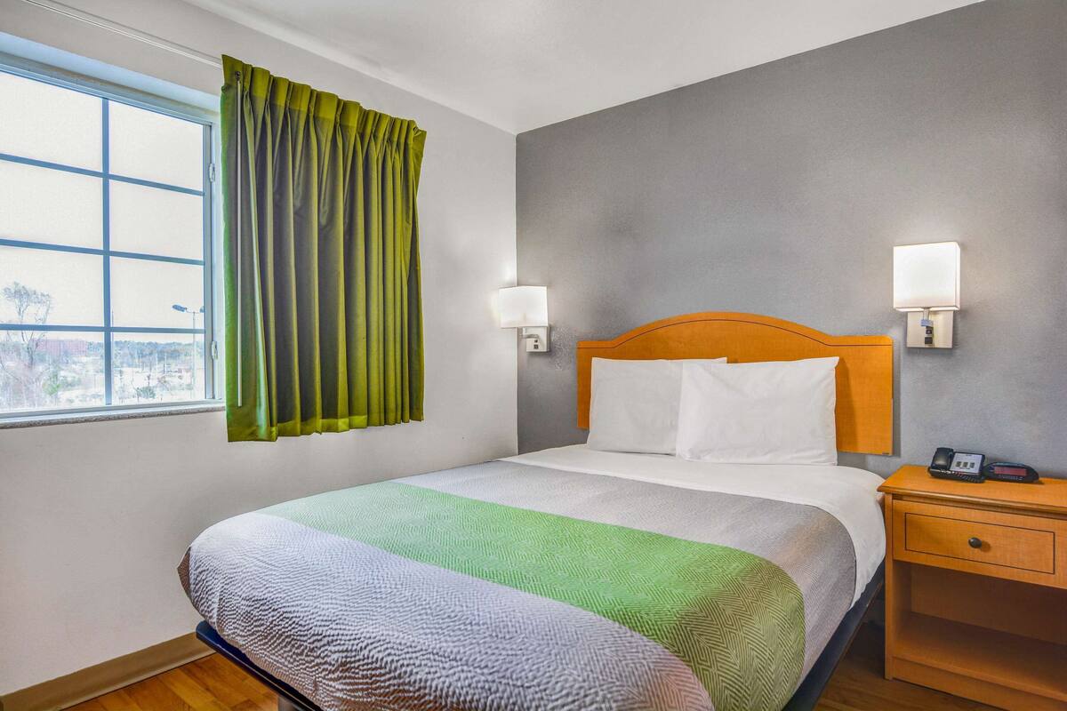 Quarto de hotel com uma cama grande, duas luminárias de cabeceira, uma mesa de cabeceira de madeira e uma janela com cortinas.