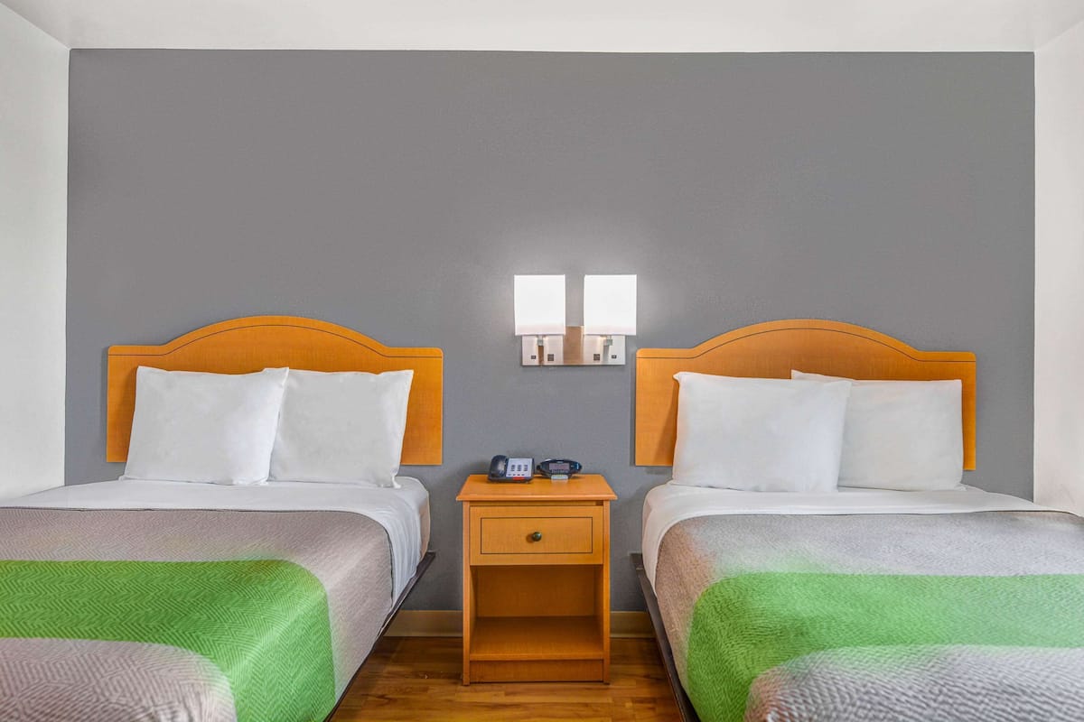 Quarto de hotel com duas camas de solteiro, uma mesa de cabeceira de madeira com telefone e uma luminária pequena, e uma parede de destaque cinza.