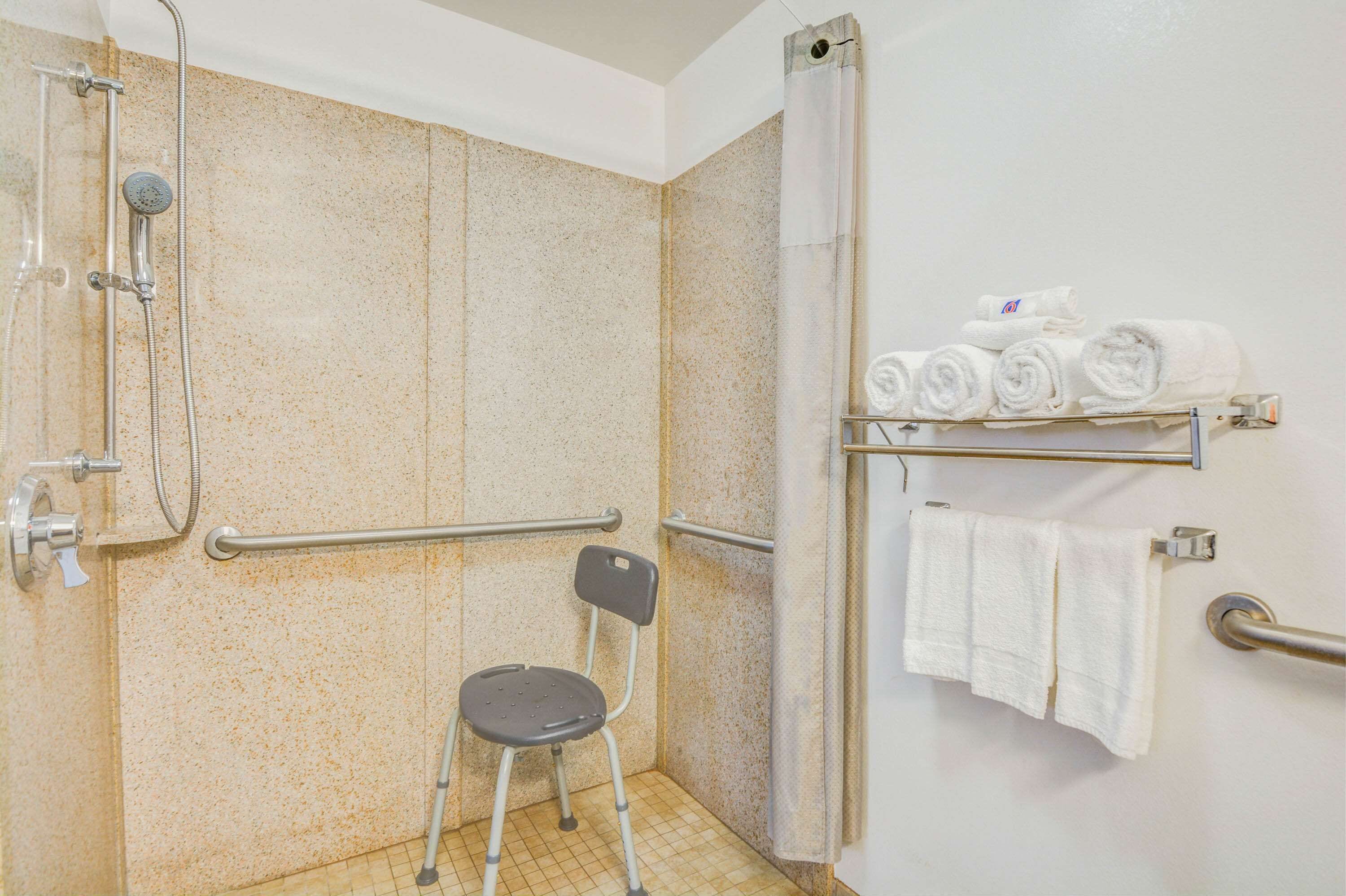 A bathroom with a shower, grab bars, a wheelchair-accessible chair, and a towel rack.