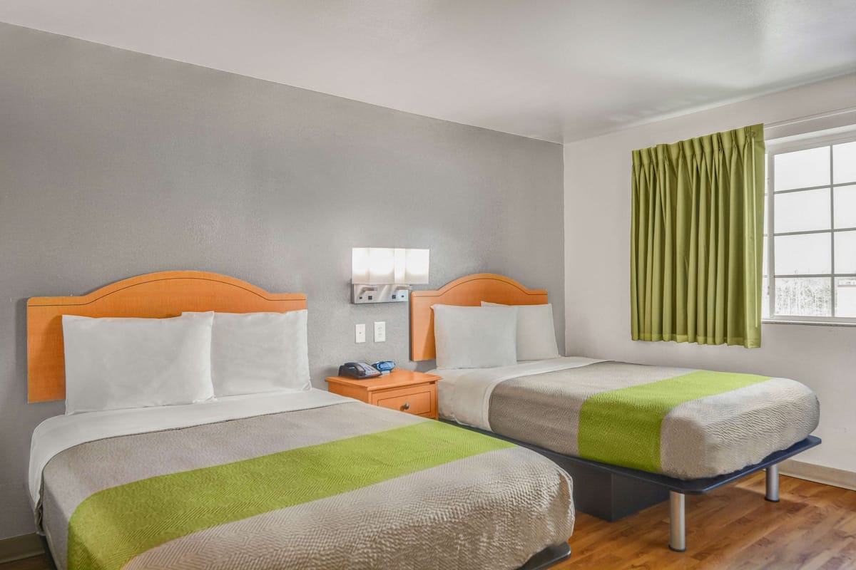 Quarto de hotel com duas camas de solteiro, uma mesa de cabeceira com telefone e uma janela com cortinas verdes.