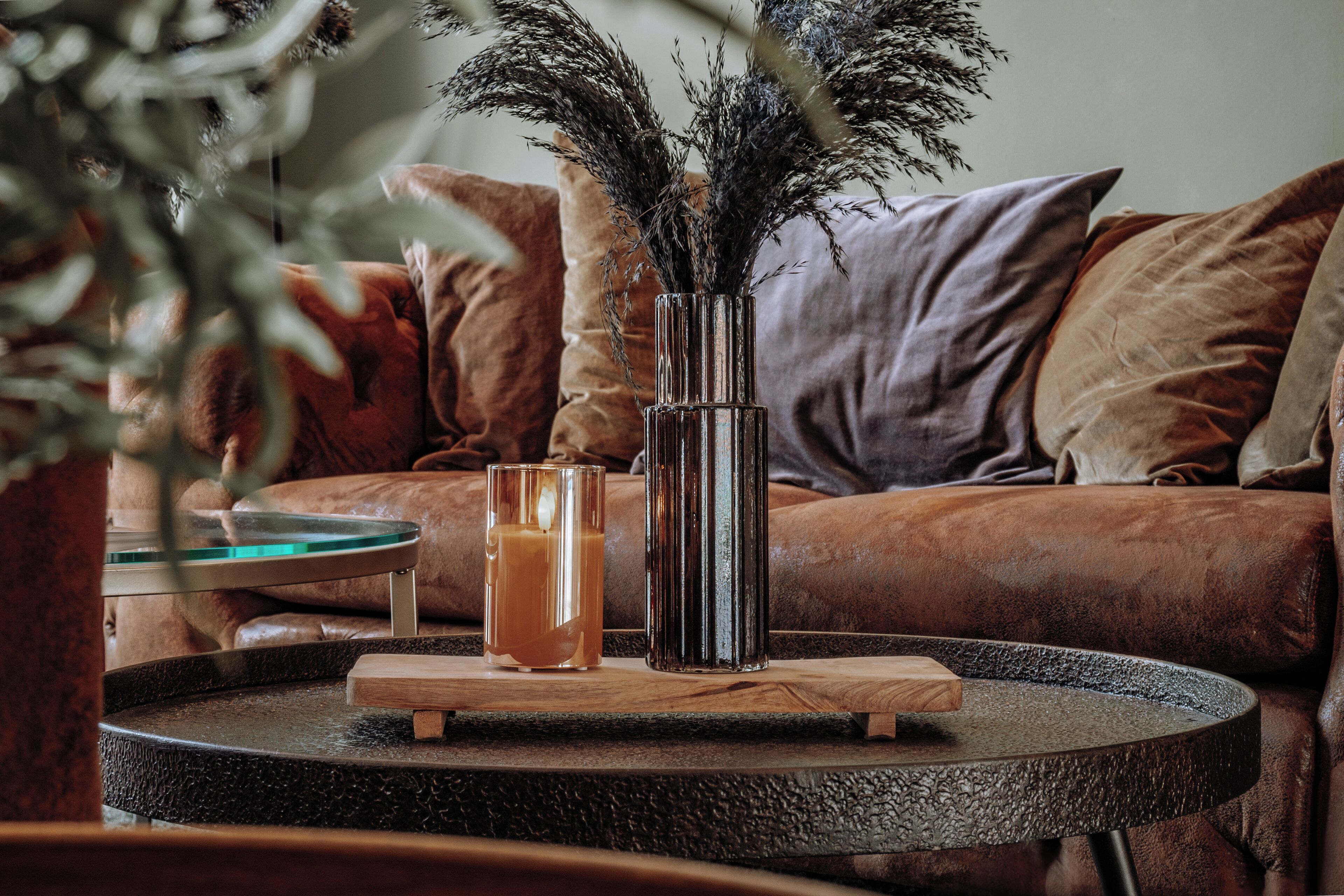 A round table with a glass and a metal vase, a wooden tray, and a brown leather sofa with cushions.
