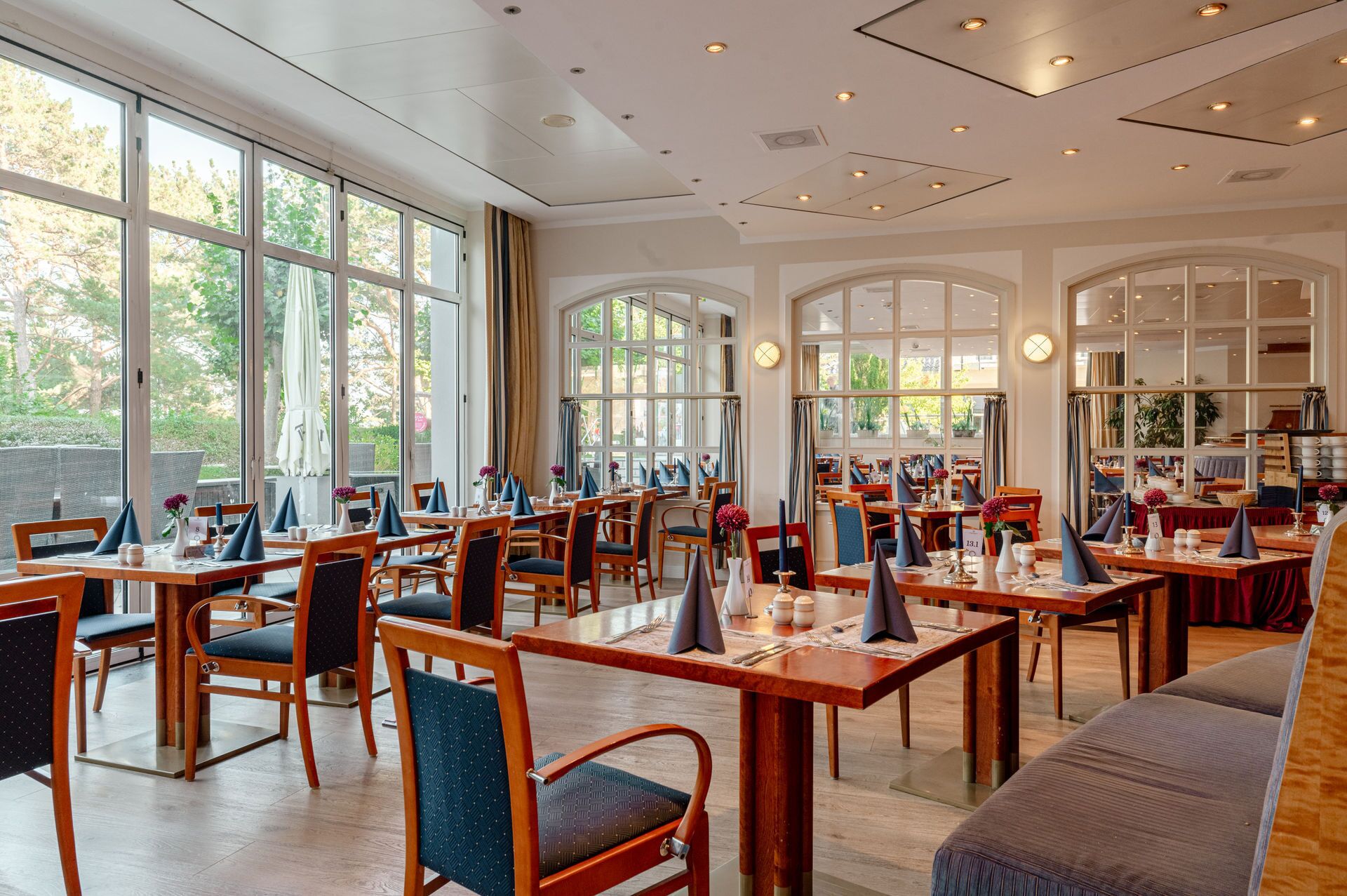 A spacious restaurant with multiple dining tables and chairs, large windows offering views of the outdoors, and a modern ceiling design.