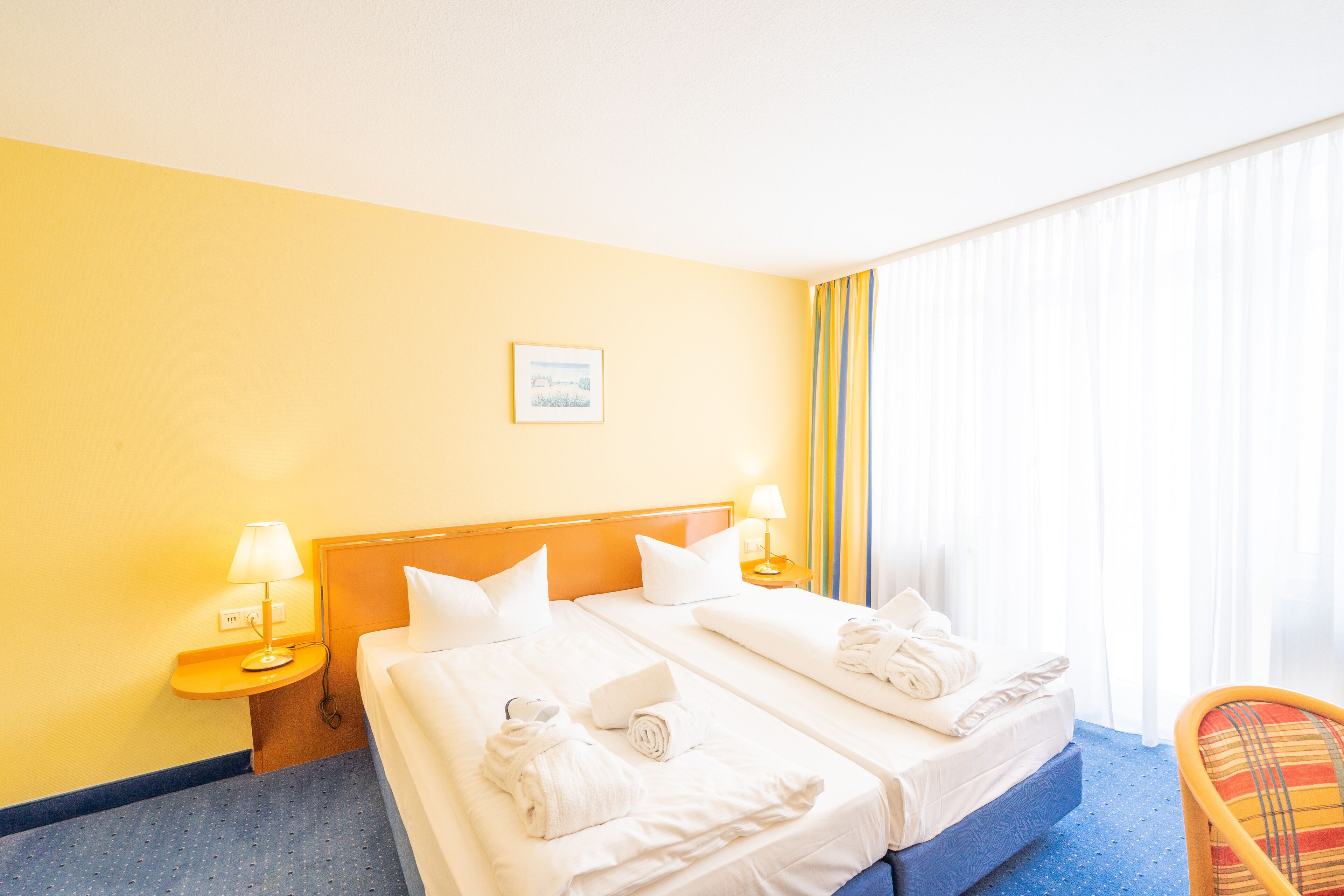 Habitación de hotel con dos camas, una mesita de noche con lámpara, una silla y una ventana con cortinas.