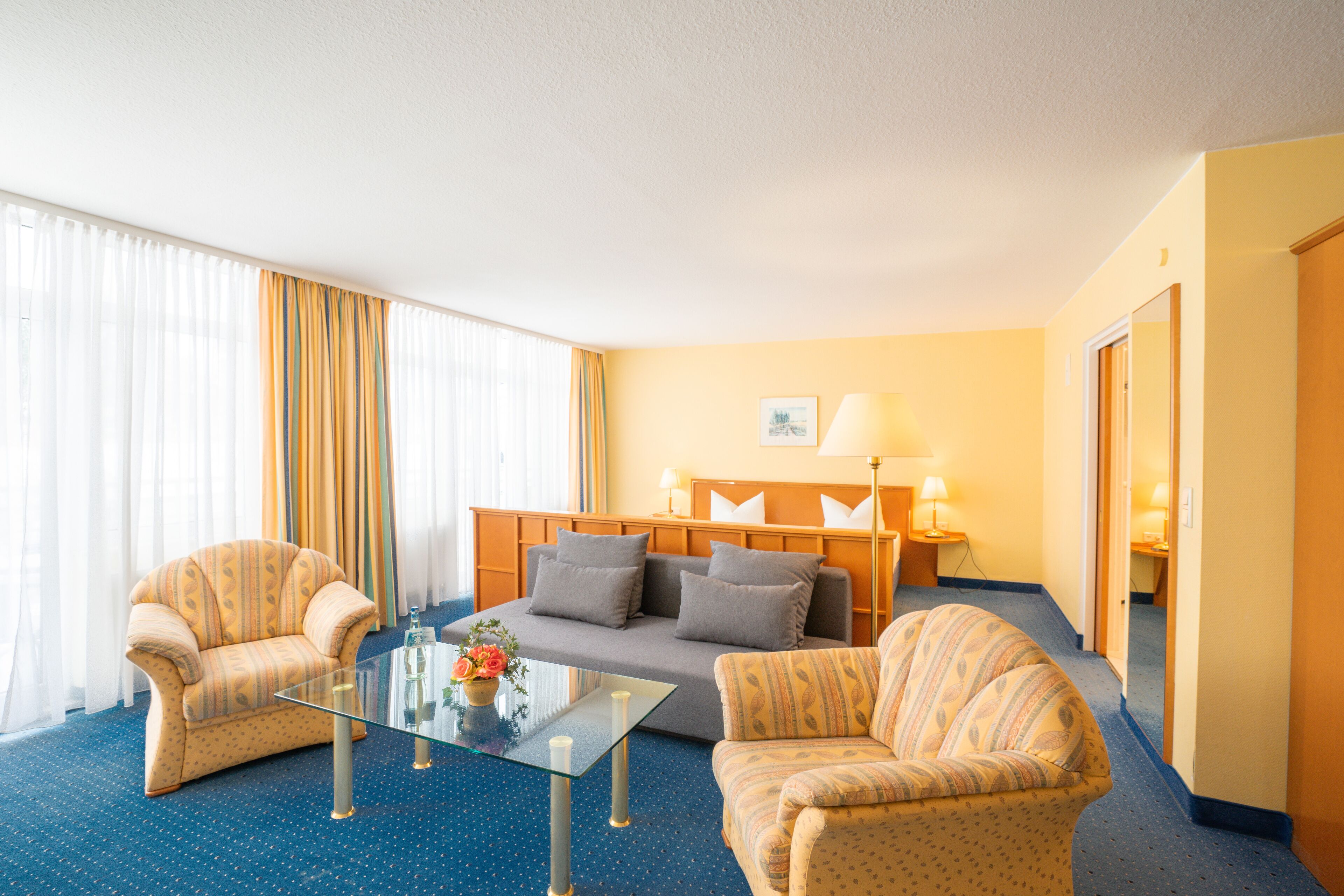A hotel room with a sofa, two armchairs, a glass coffee table, and a lamp.