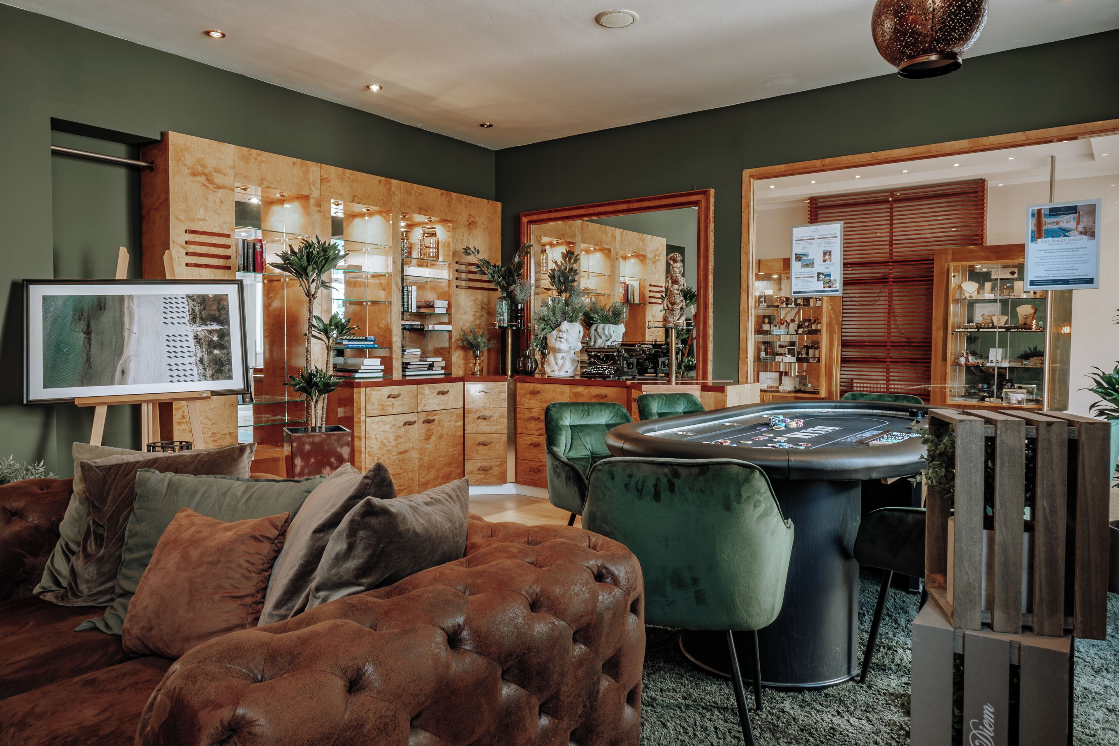 A modern living room with a tufted brown sofa, a green velvet armchair, and a black poker table. There is a wooden cabinet with glass doors displaying various items.